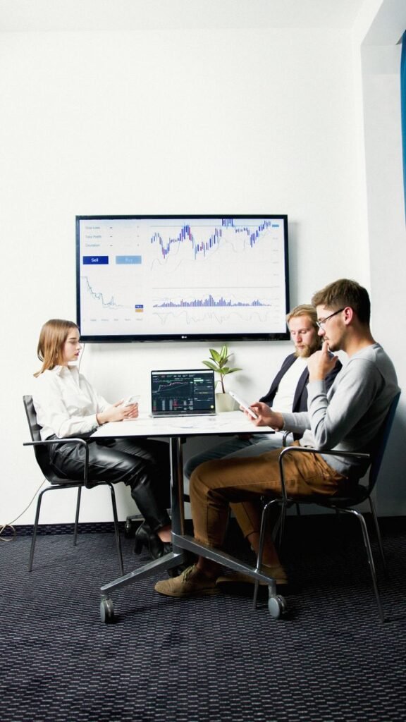 pexels-photo-5715850-5715850 Three business professionals in a meeting room discussing financial charts displayed on a screen.